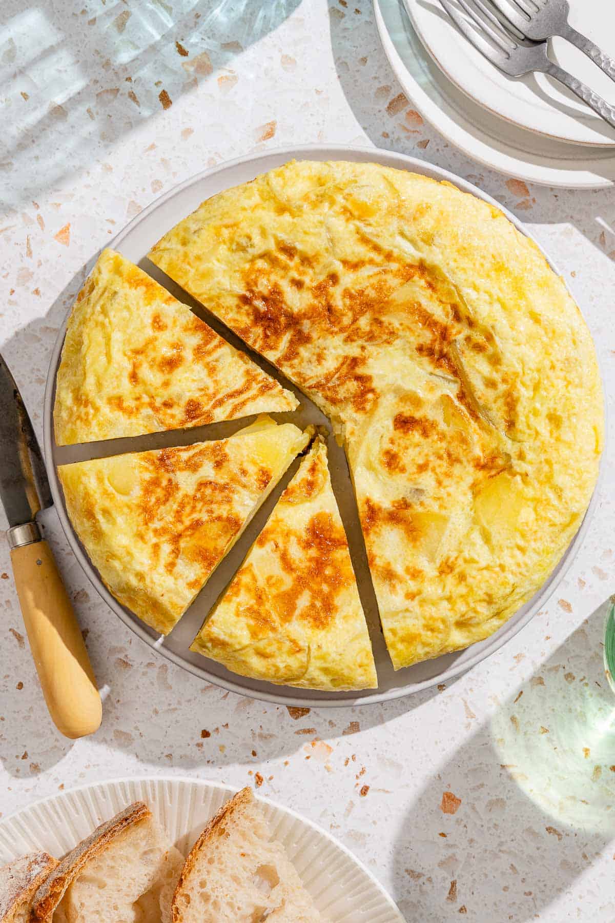 The partially sliced spanish tortilla on a platter. Next to this is a stack of 2 plates with 2 forks, a knife, and a plate with sliced bread.