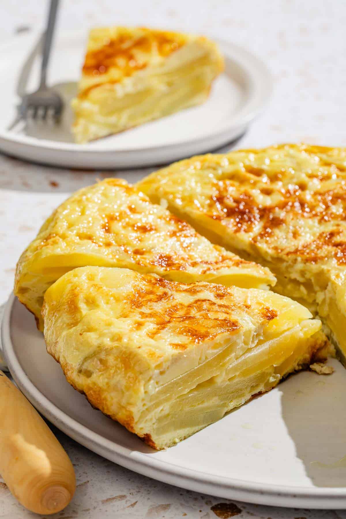 The partially sliced spanish tortilla on a platter with one sliced removed. Next to this is a slice of the tortilla on a plate with a fork.