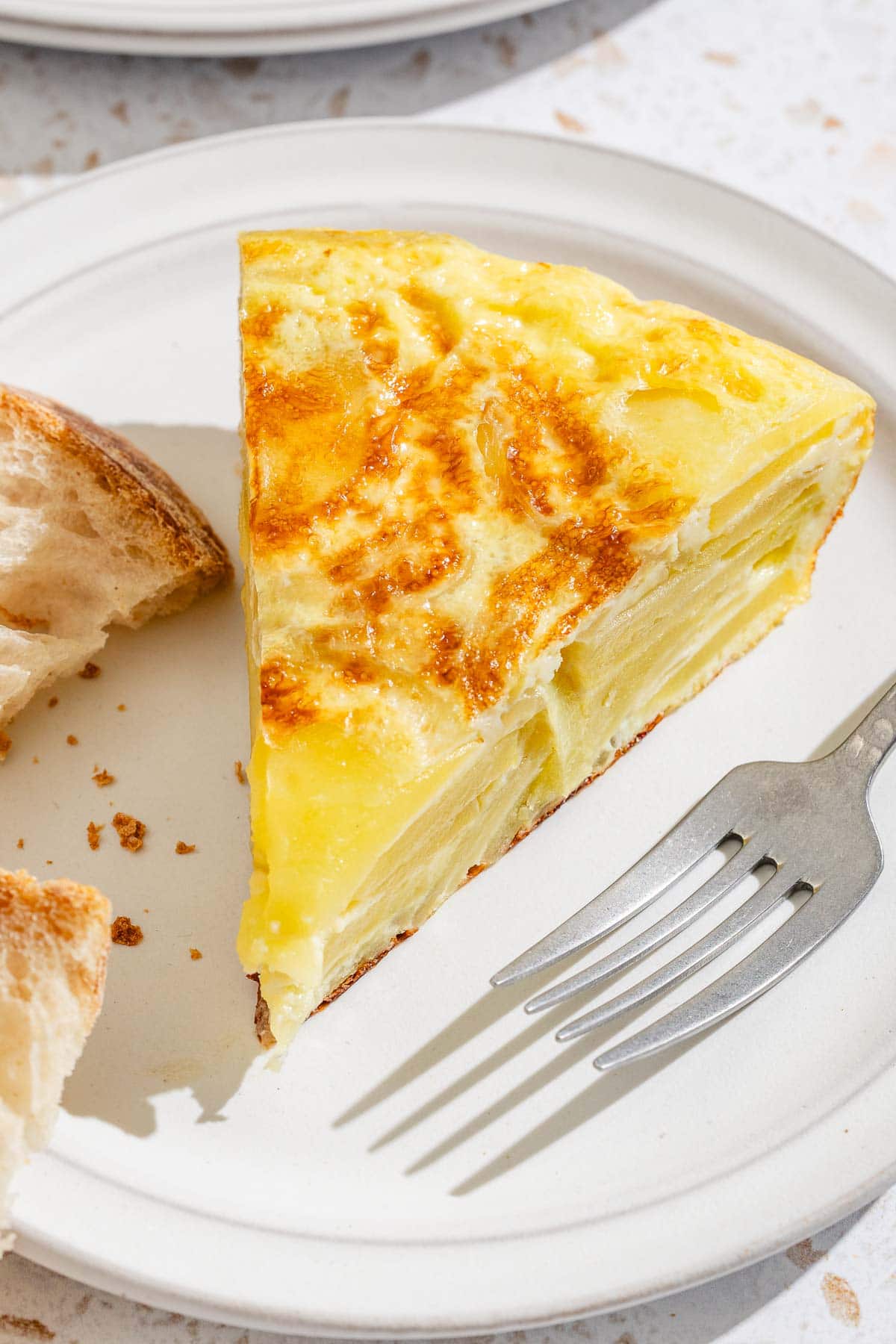 A slice of the spanish tortilla on a plate with a slice of bread and a fork.