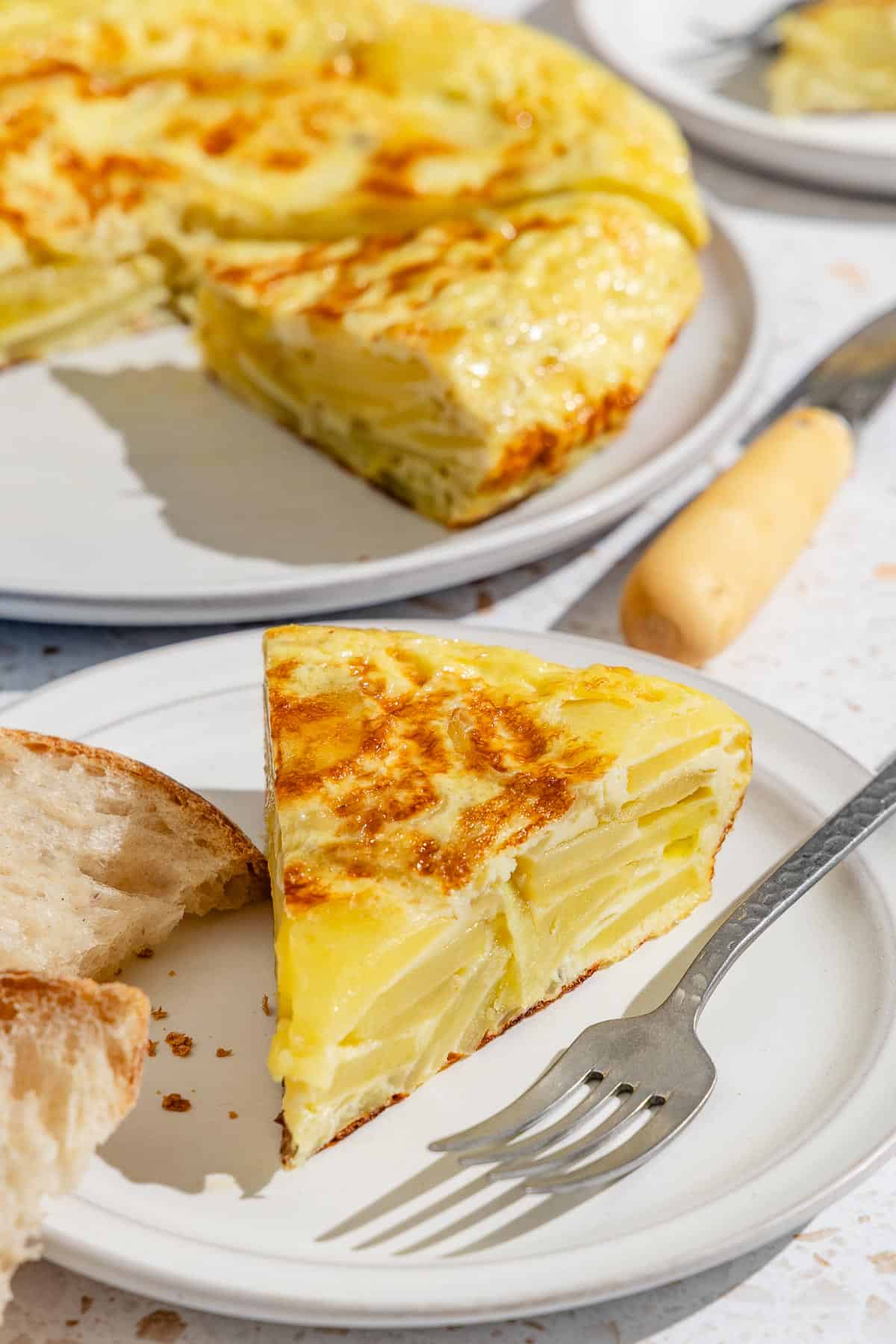 A slice of the spanish tortilla (Tortilla de Patatas) on a white plate with a fork. In the background is the rest of the tortilla on a platter and a knife.