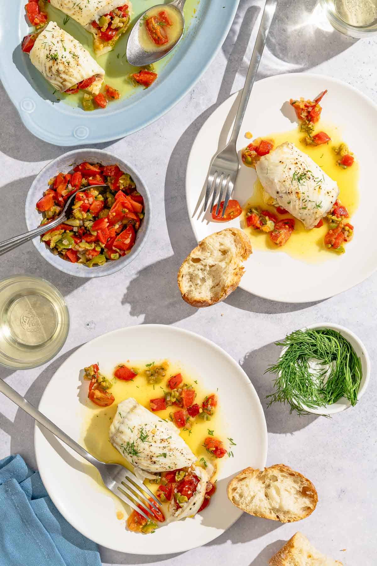 Two servings of the stuffed flounder each on its own plate along with a fork and a slice of crusty bread. Next to these is a bowl of fresh dill, a cloth napkin, a both of the stuffing with a spoon, a glass of white wine, and the rest of the stuffed flounder on a platter with a spoon.