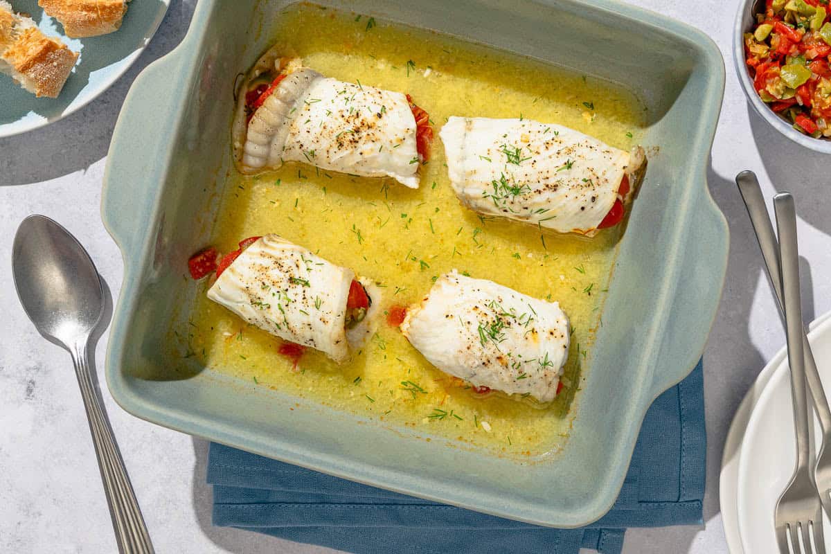 Four stuffed flounder fillets in a baking dish with chicken stock, white wine, garlic and saffron. Next to this is a serving spoon, a stack of 2 plates with 2 forks, a bowl of the stuffing, and a plate with slices of crusty bread.