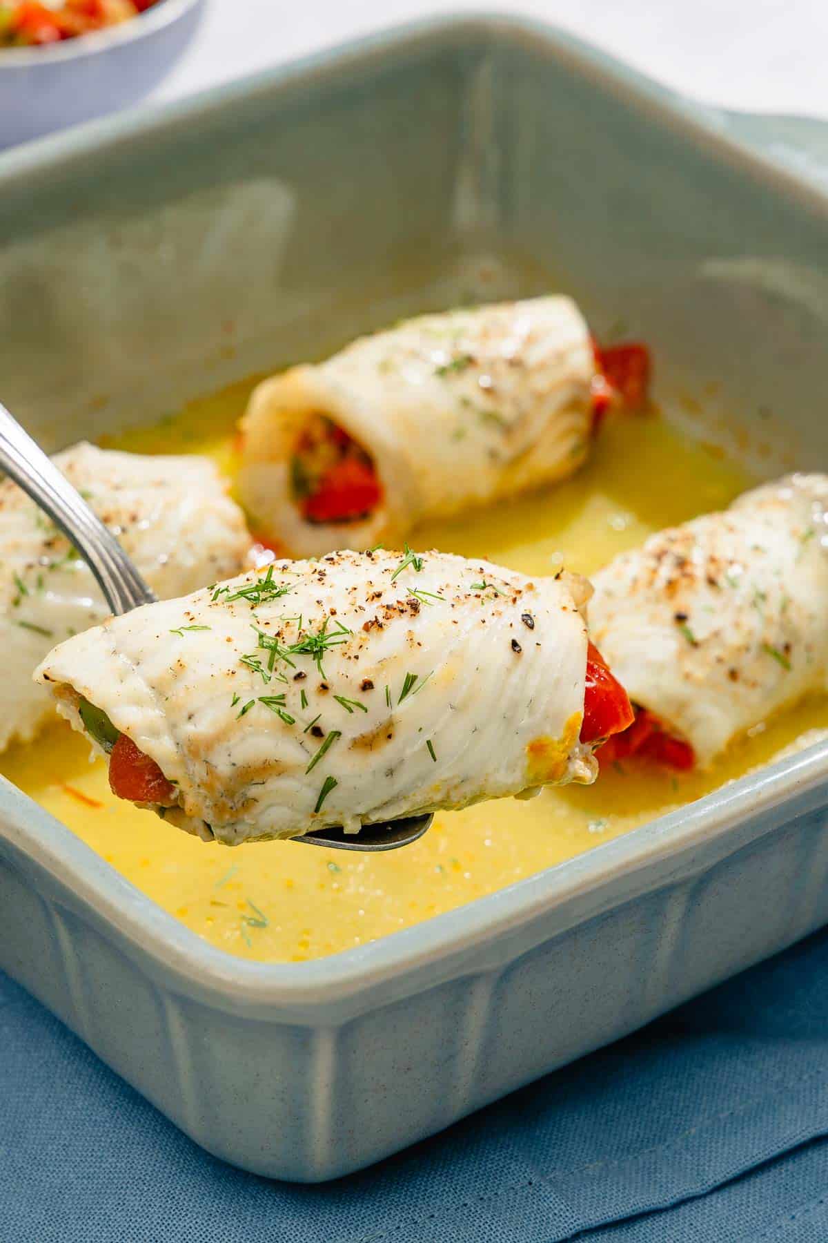 A serving spoon holding 1 stuffed flounder fillet being held over a backing dish with the other 3 stuffed flounder fillets.