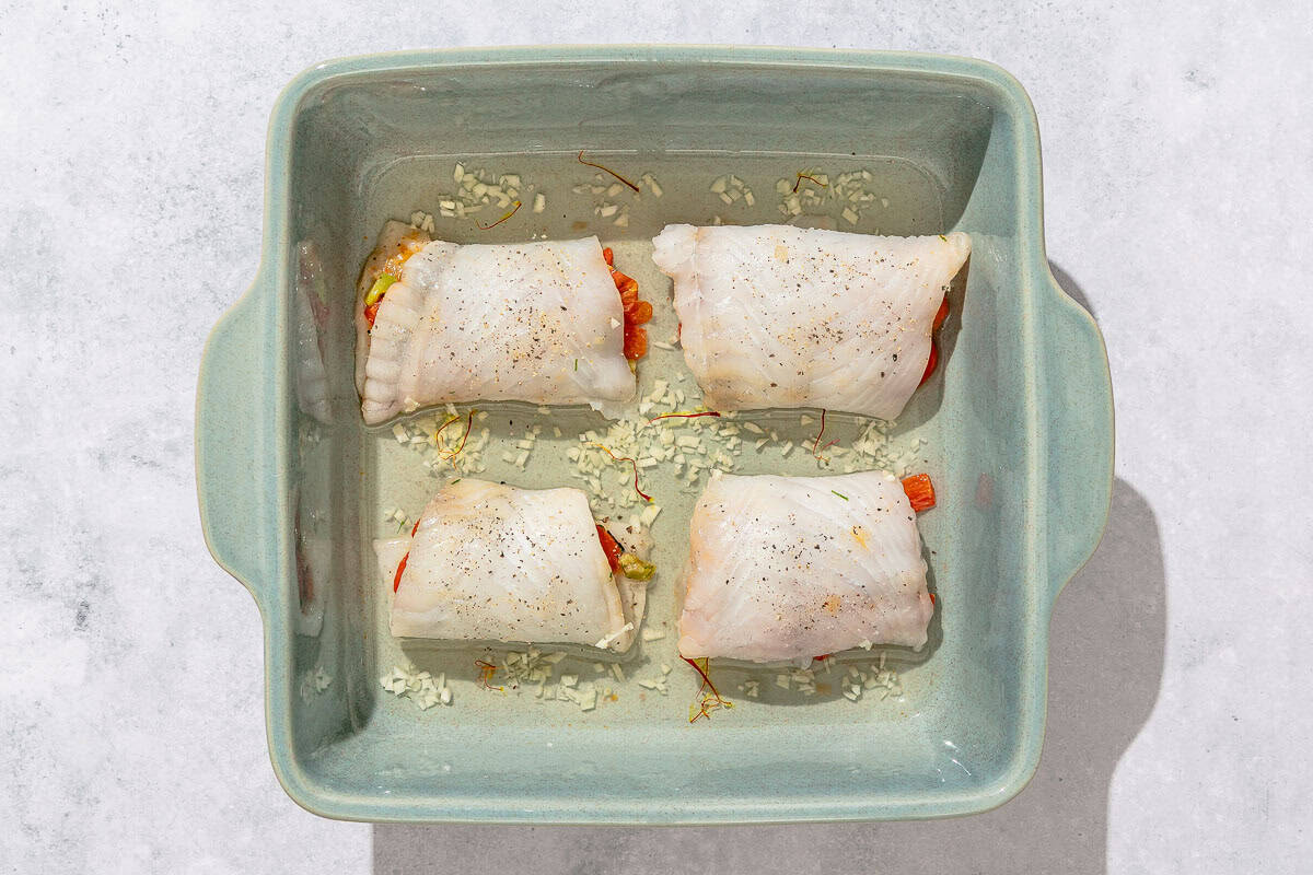 Four uncooked stuffed flounder fillets in a baking dish with chicken stock, white wine, garlic and saffron.
