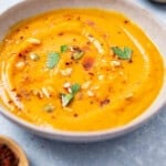 A close up of a bowl of butternut squash apple soup next to an empty bowl and a bowl of aleppo pepper.