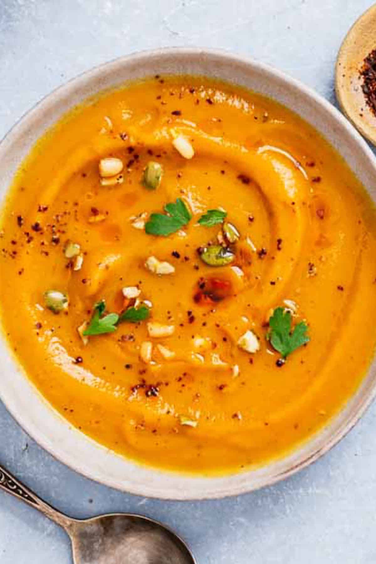 An overhead photo of a bowl of butternut squash apple soup next to a spoon and a bowl of aleppo pepper.