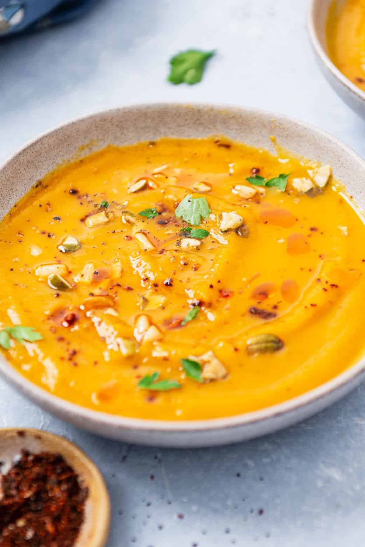 A close up of a bowl of butternut squash apple soup next to a bowl of aleppo pepper.