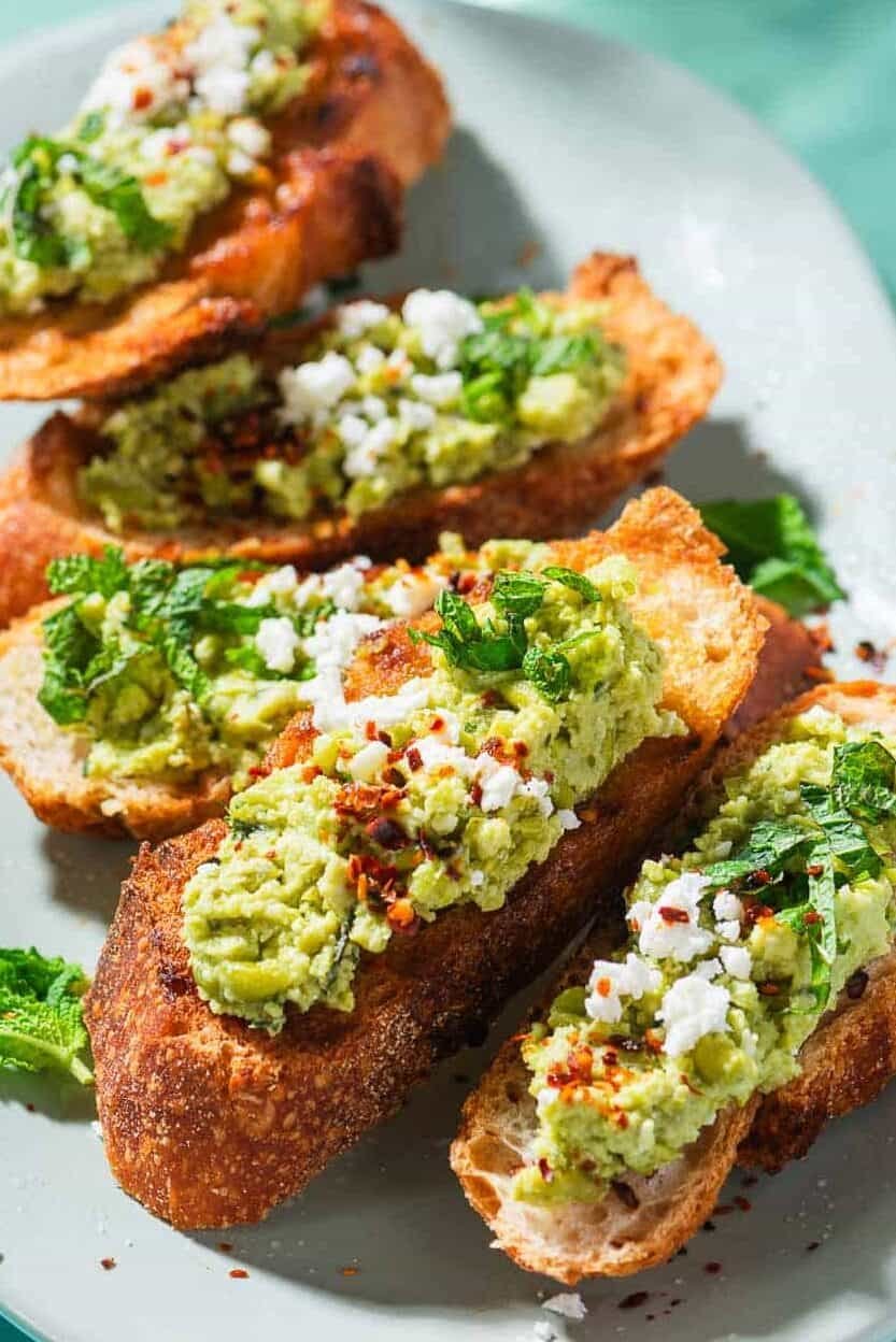A close up photo of 4 pieces of smashed peas on toast garnished with aleppo pepper, feta and mint on a serving platter. Next to this is a bowl of the smashed peas, a small bowl of Aleppo pepper, and3 pieces of tasted bread.