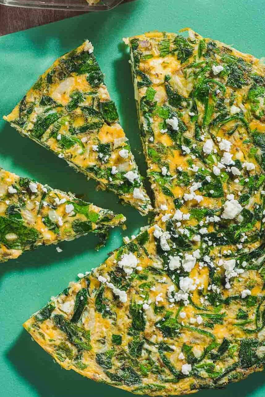 An overhead photo of a feta spinach frittata on a cutting board with two slices cut from it. Next to this is a mediterranean cucumber salad in a bowl with a spoon.