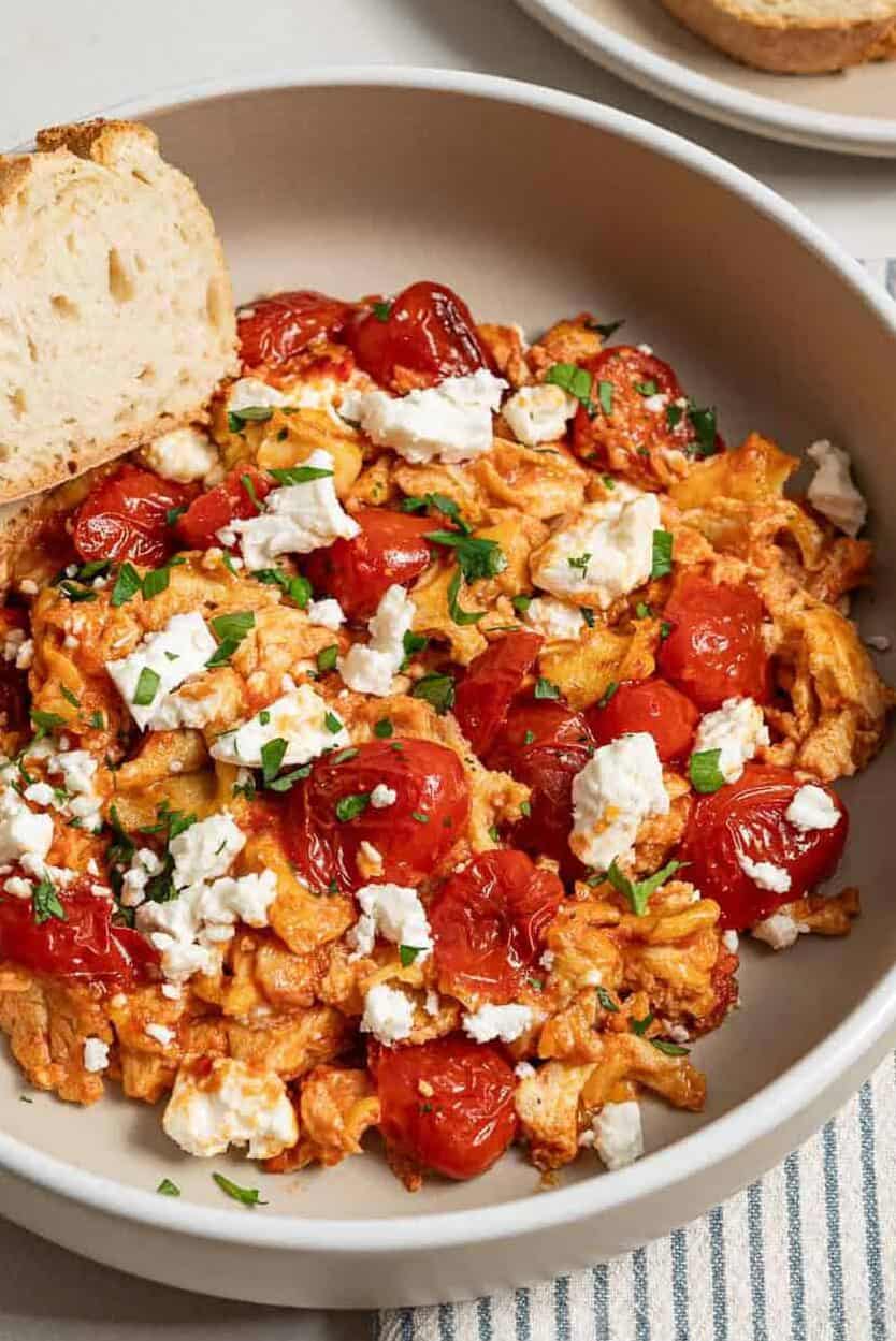 A close up of a serving of strapastada greek tomato scrambled eggs topped with crumbled feta and chopped parsley in a bowl with 2 slices of bread. Next to this is a cloth napkin and a plate of sliced bread.