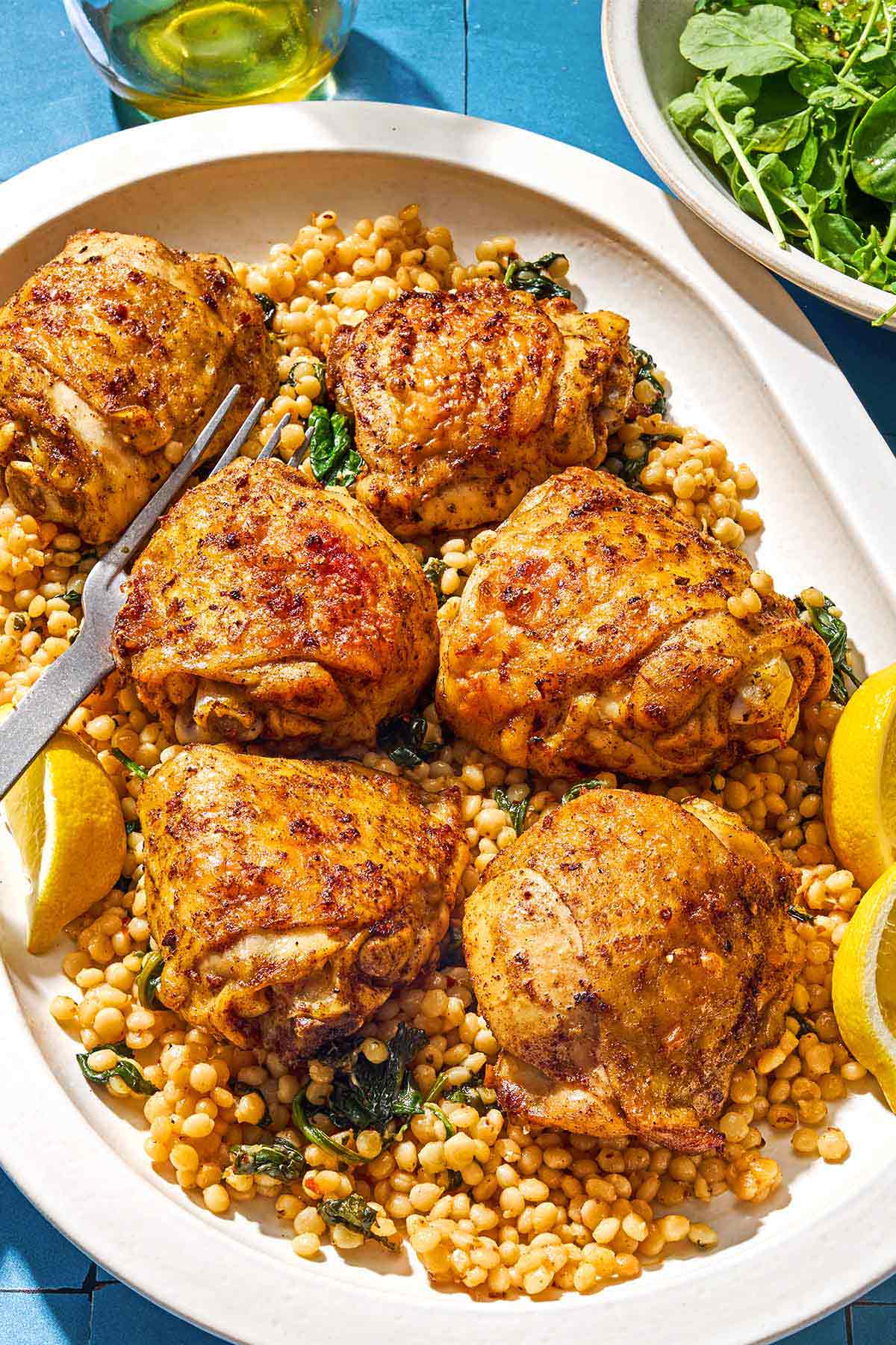 Six air fryer chicken thighs on a bed of spinach couscous on a platter with a fork. Next to this is a bowl of salad and a bottle of olive oil.