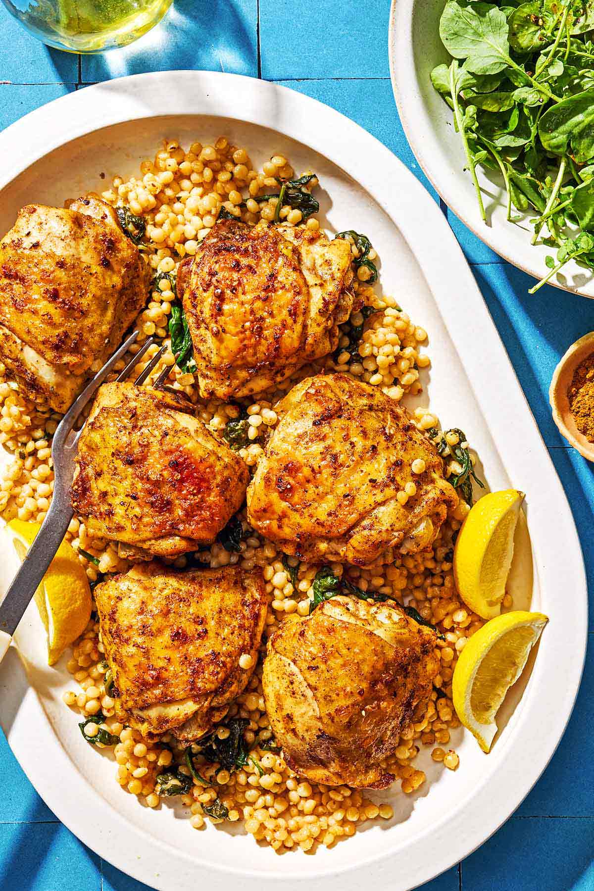 Six air fryer chicken thighs on a bed of spinach couscous on a platter with a fork and lemon wedges. Next to this is a bowl of salad, a bowl of ras el hanout, and a bottle of olive oil.