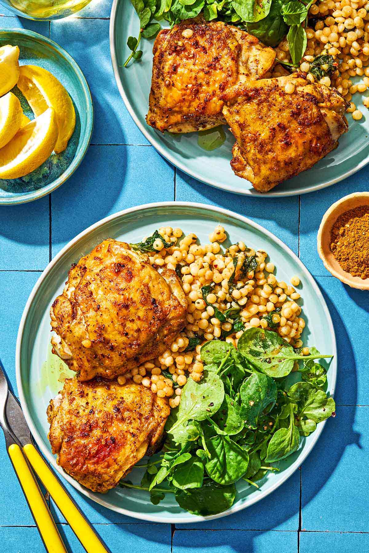 An overhead photo of 2 plates of air fryer chicken, salad and spinach couscous. Next to these are bowls of ras el hanout and lemon wedges and a knife and fork.