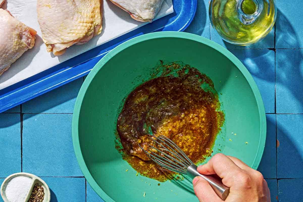 Marinade for the air fryer chicken thighs being stirred in a bowl with a whisk. Next to this is a bottle of olive oil, a small bowl of salt and pepper, and uncooked chicken thighs on a parchment lined sheet pan.