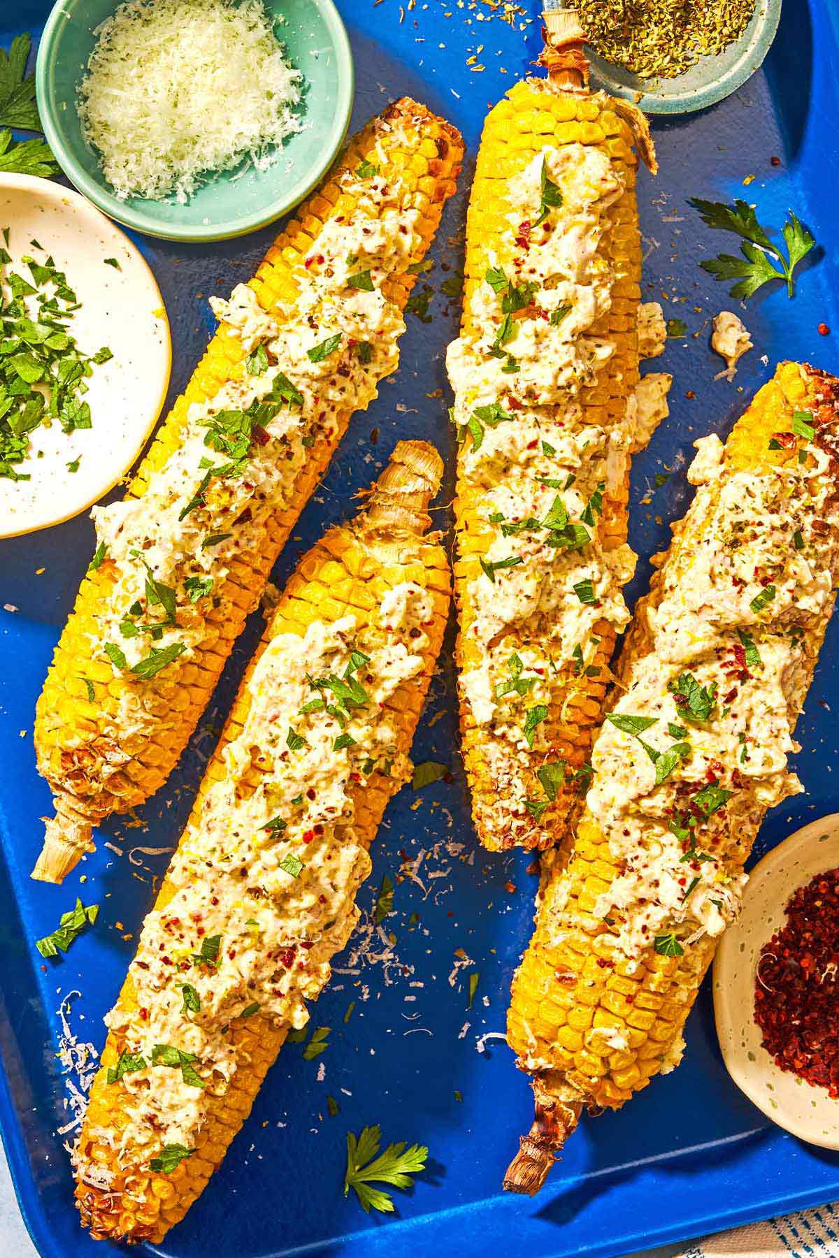 Four ears of air fryer corn on the cob topped with the feta-yogurt sauce, chopped parsley, aleppo pepper and dried oregano on a sheet pan with bowls of chopped parsley, grated parmesan cheese, dried oregano and aleppo pepper.