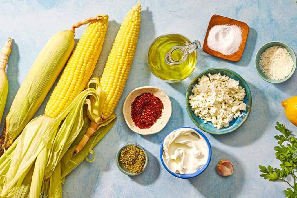 Ingredients for air fryer corn on the cob including ears of corn, olive oil, salt, aleppo pepper, parsley, garlic, feta, Greek yogurt, dried oregano, grated parmesan cheese and lemon.