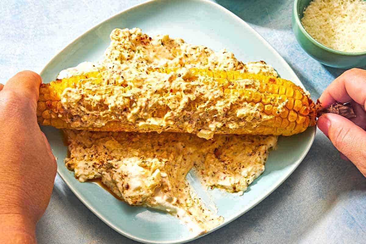 An ear of air fryer corn being rolled in the feta sauce on a plate. Next to this is a bowl of grated parmesan cheese.