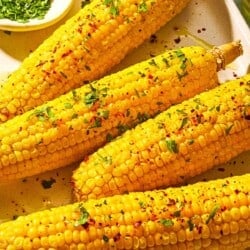 Four ears of air fryer corn on the cob topped with a drizzle of olive oil, aleppo pepper, chopped parsley and lemon zest on a sheet pan with small bowls of chopped parsley and aleppo pepper.