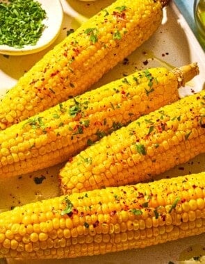 Four ears of air fryer corn on the cob topped with a drizzle of olive oil, aleppo pepper, chopped parsley and lemon zest on a sheet pan with small bowls of chopped parsley and aleppo pepper.