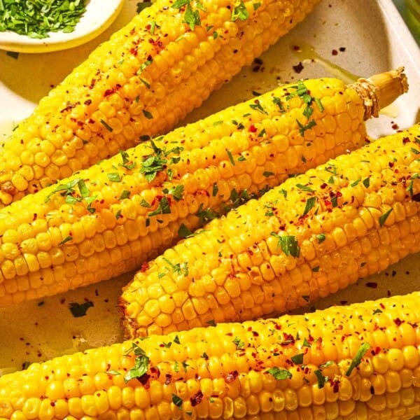 Four ears of air fryer corn on the cob topped with a drizzle of olive oil, aleppo pepper, chopped parsley and lemon zest on a sheet pan with small bowls of chopped parsley and aleppo pepper.