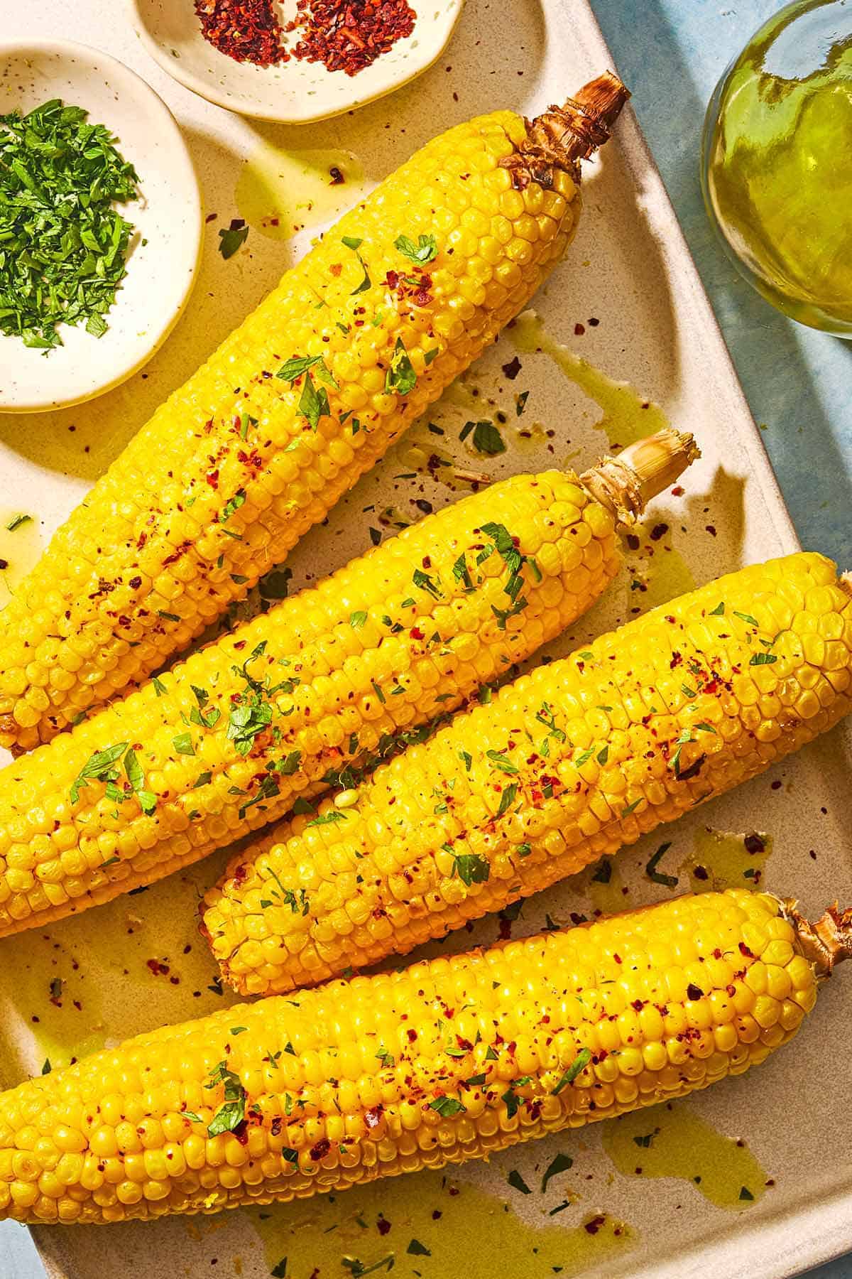 Four ears of air fryer corn on the cob topped with a drizzle of olive oil, aleppo pepper, chopped parsley and lemon zest on a sheet pan with small bowls of chopped parsley and aleppo pepper. Next to the sheet pan is a bottle of olive oil.