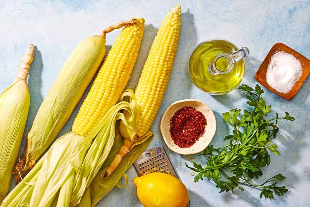 Ingredients for air fryer corn on the cob including ears of corn, olive oil, salt, aleppo pepper, parsley, and lemon.