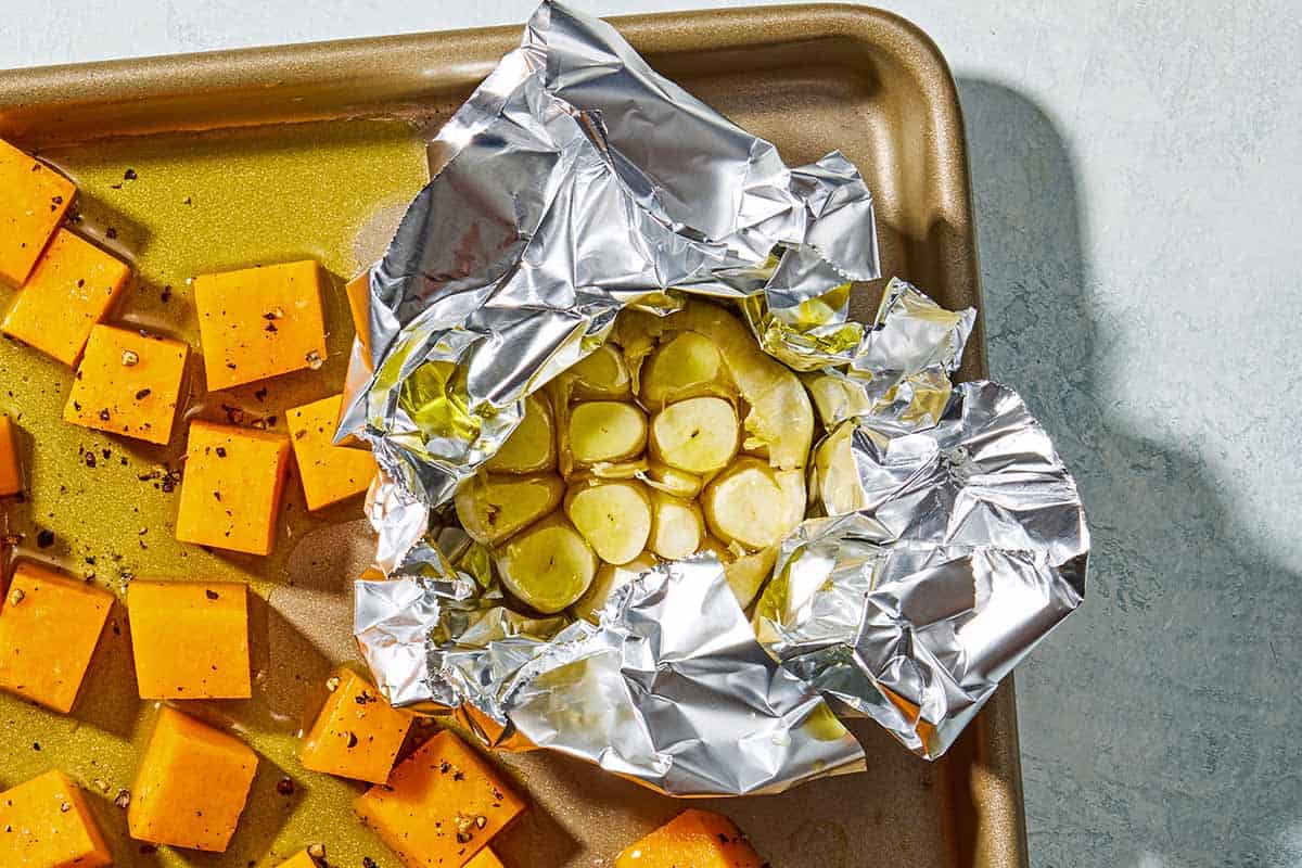 A head of garlic drizzled with olive oil and wrapped in foil on a sheet pan with cubes of seasoned butternut squash.