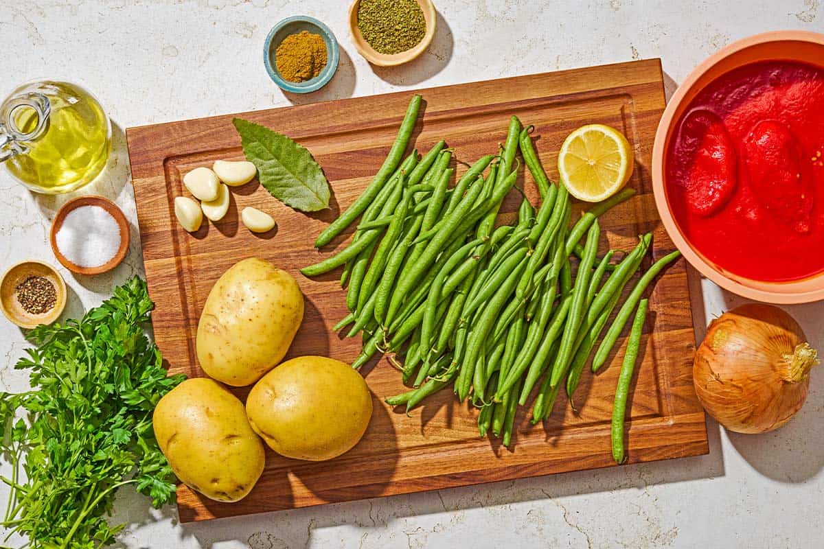 Ingredients for fasolakia including green beans, olive oil, onion, garlic, ground cumin, dried oregano, canned peeled whole tomatoes, a bay leaf, potatoes, salt, black pepper, lemon and parsley.