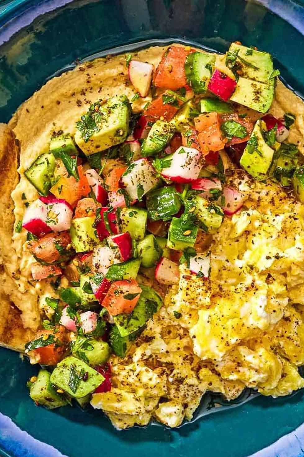 A bowl of breakfast hummus bowl with scrambled eggs and cucumber tomato salad on top.