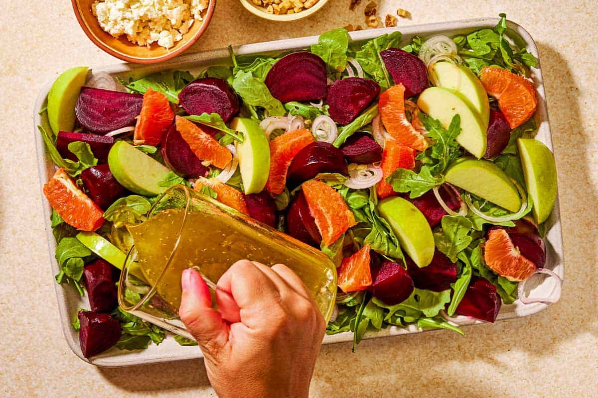 The ladolemono dressing being poured over the beet salad on a platter. Next to this is a bowl of crumbled feta, and a bowl of chopped walnuts.