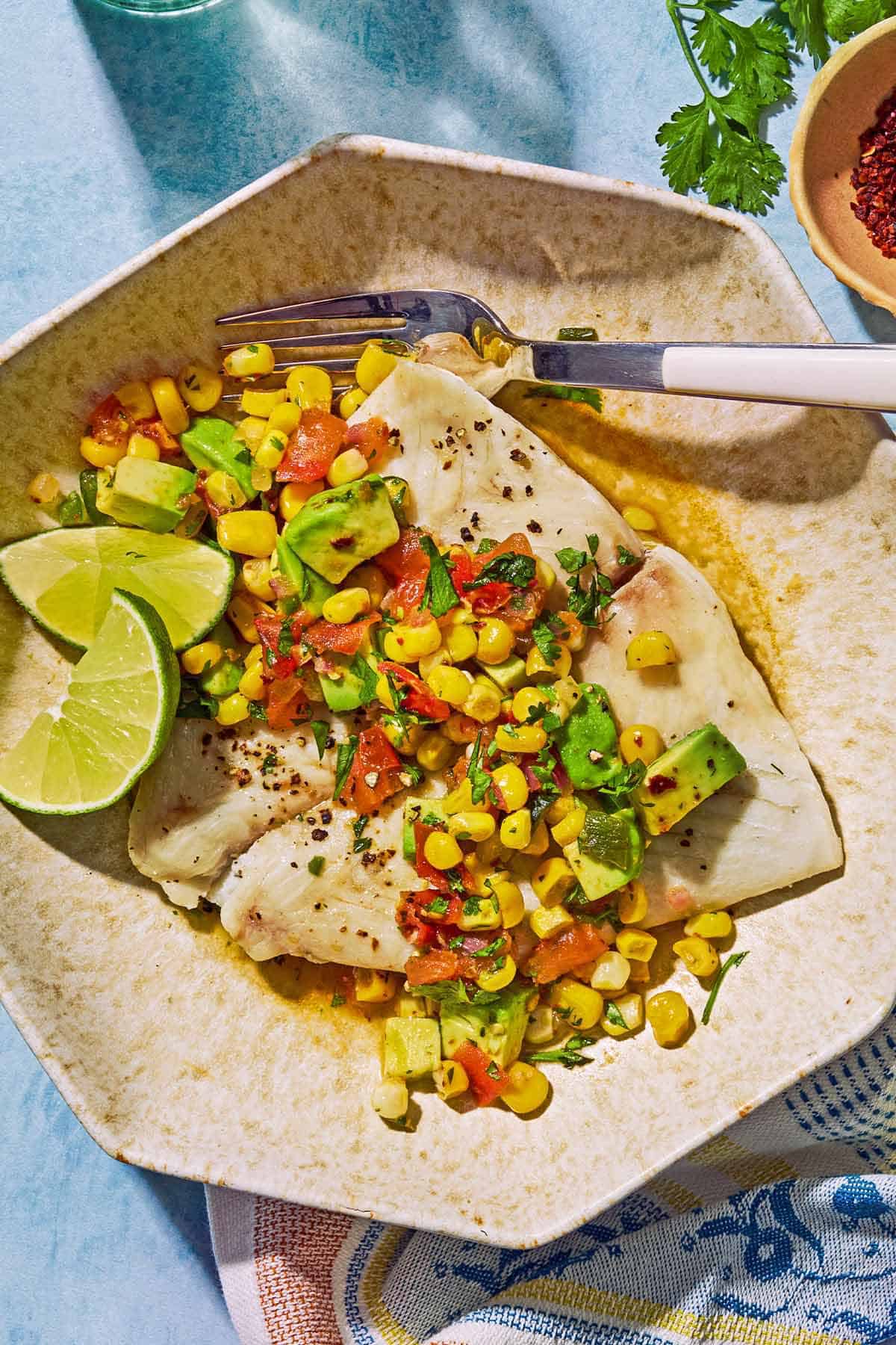 Poached flounder topped with sweet corn salsa on a plate with lime wedges and a fork. Next to this is a kitchen towel, a bowl of aleppo pepper and some sprigs of cilantro.
