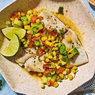 Poached flounder topped with sweet corn salsa on a plate with lime wedges.