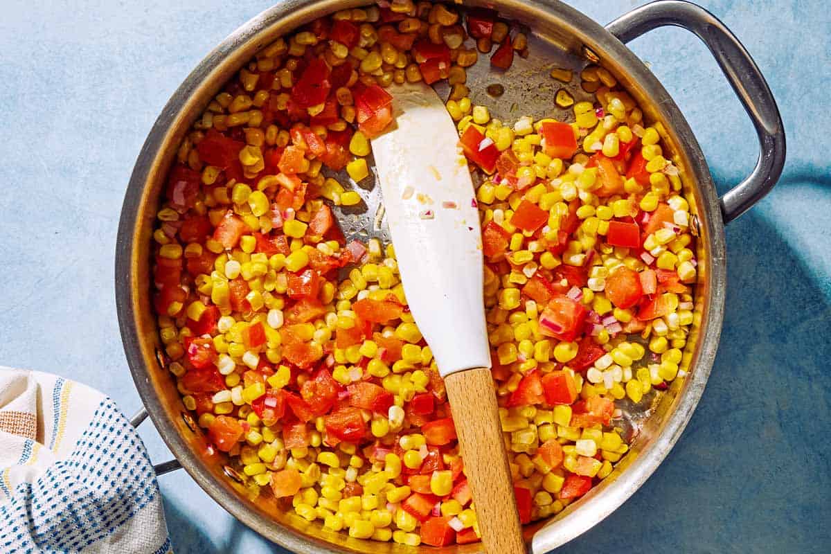 The corn, tomato, onion, and garlic for the sweet corn salsa being cooked in a skillet with a spatula.