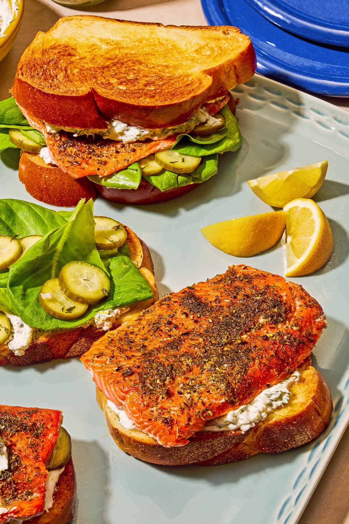 A salmon sandwich, lemon wedges, and a open-faced salmon sandwich next to its top half on a platter.