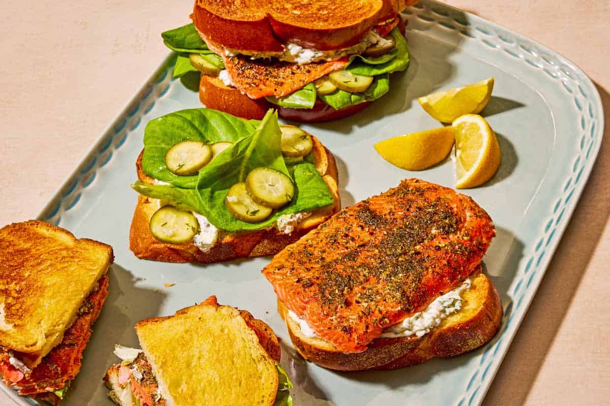 Three salmon sandwiches on a platter with lemon wedges. One is whole, one is cut in half, and one is open-faced sitting next to it's top half.