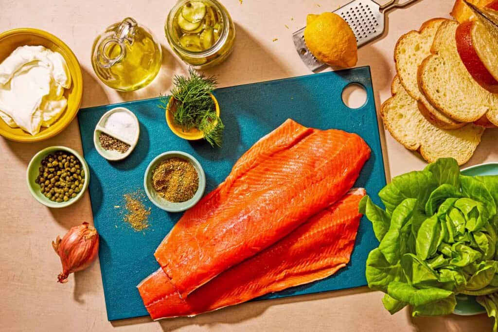 Ingredients for salmon sandwich including salmon, olive oil, seafood seasoning, salt, black pepper, butter lettuce, quick pickled cucumbers, challah bread, greek yogurt, shallots, fresh dill, capers, and lemon.