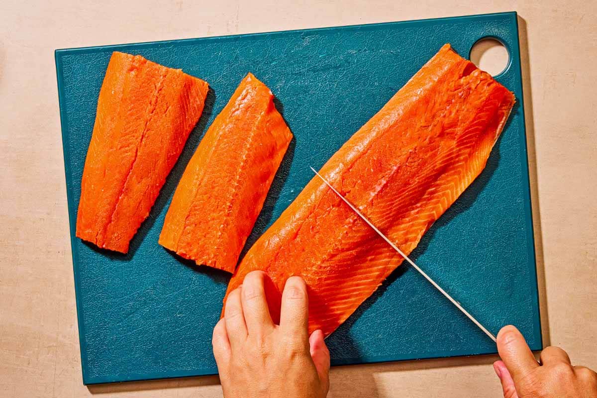 Two salmon fillets and one larger piece of salmon being cut in half on a cutting board.