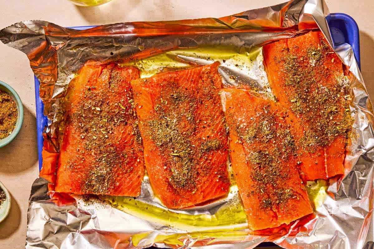 Four seasoned uncooked salmon fillets on a foil lined baking sheet.