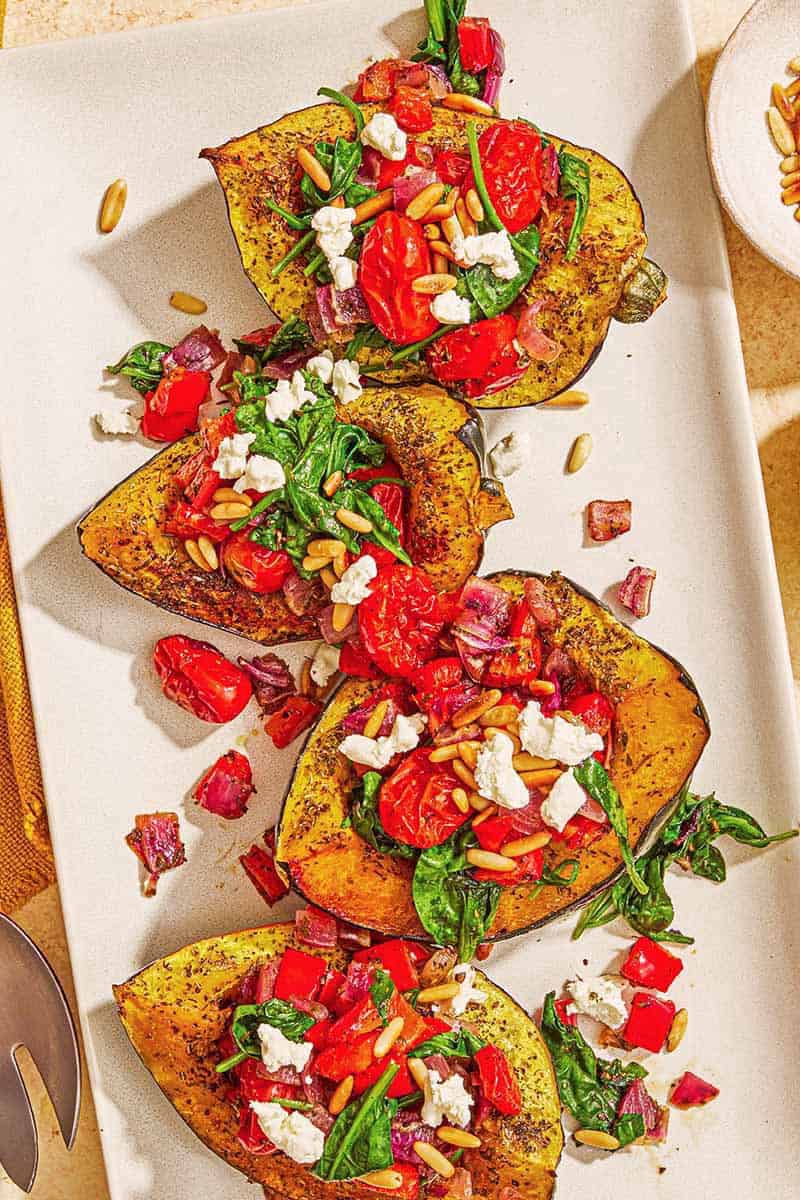 Four stuffed acorn squash halves on a white platter filled with tomatoes, spinach and goat cheese.