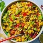 Sweet corn salsa in a bowl with a spoon. Next to this is a kitchen towel, a bowl of aleppo pepper, a sprig of cilantro and two lime halves.