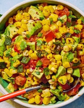 Sweet corn salsa in a bowl with a spoon. Next to this is a kitchen towel, a bowl of aleppo pepper, a sprig of cilantro and two lime halves.
