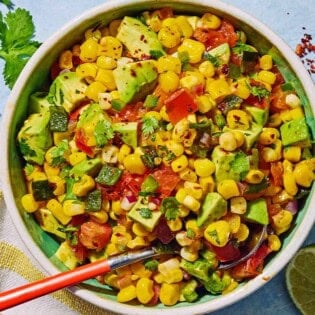 Sweet corn salsa in a bowl with a spoon. Next to this is a kitchen towel, a bowl of aleppo pepper, a sprig of cilantro and two lime halves.