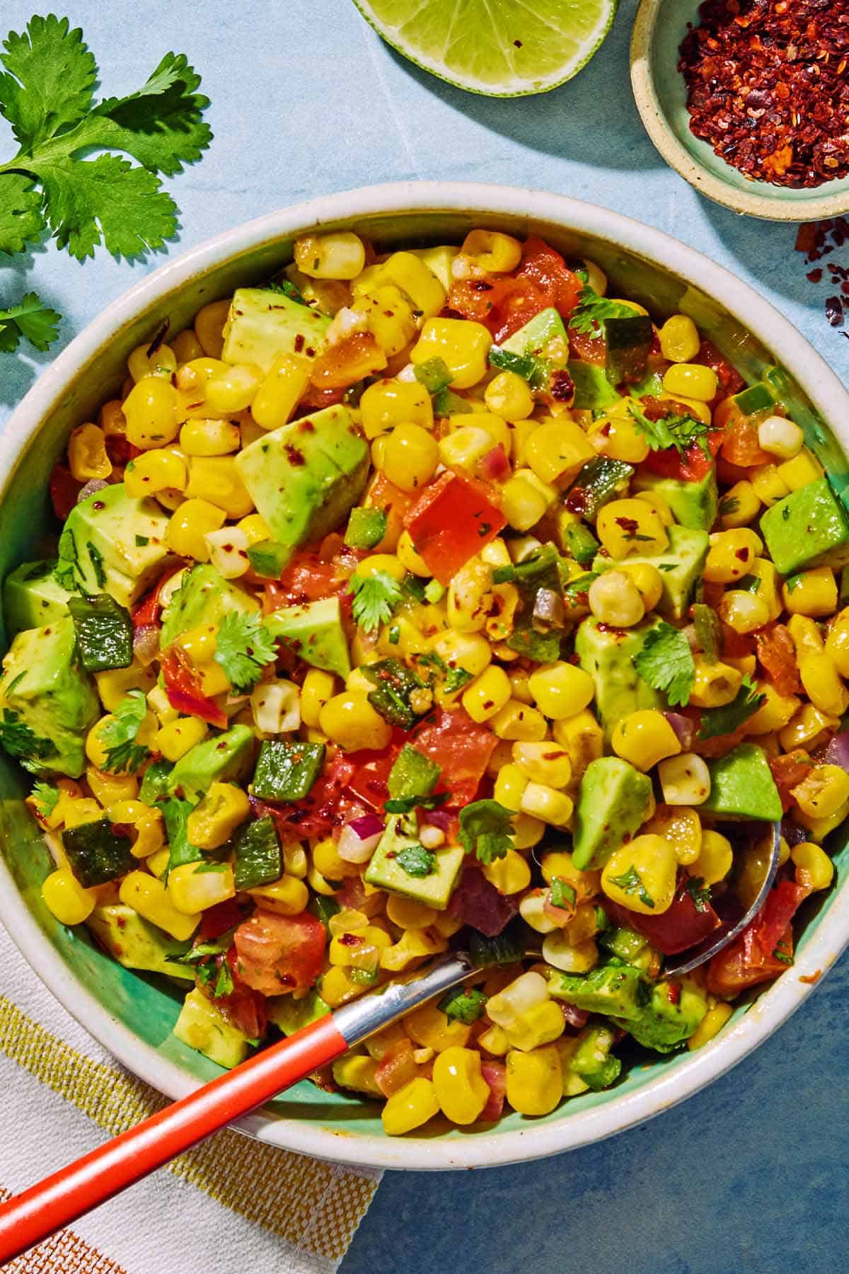Sweet corn salsa in a bowl with a spoon. Next to this is a kitchen towel, a bowl of aleppo pepper, a sprig of cilantro and a lime half.