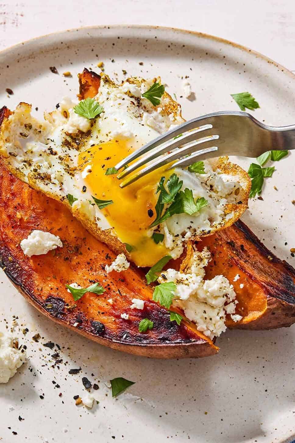 A close up of a sweet potato topped with a fried egg, spices, feta and parsley on a plate with a fork.