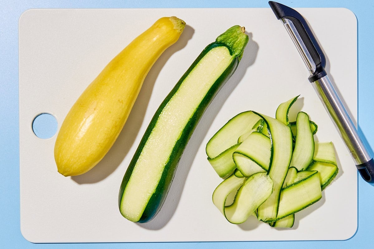 A summer squash, a partially peeled zucchini, a pile of zucchini ribbons and a vegetable peeler on a cutting board.