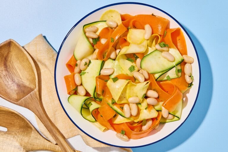 Zucchini ribbon salad in a bowl next to a cloth napkin and wooden serving utensils.