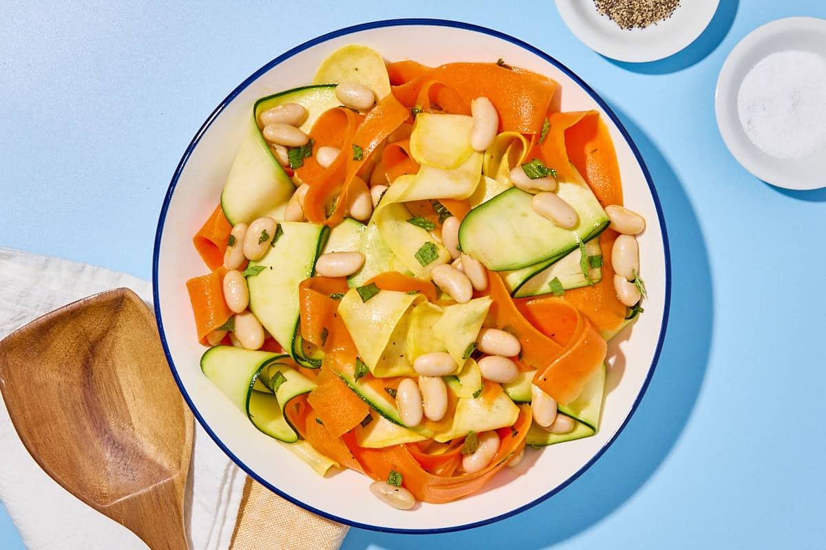 Zucchini ribbon salad in a bowl next to a cloth napkin, a wooden spoon, a glass of water and small bowls of salt and black pepper.
