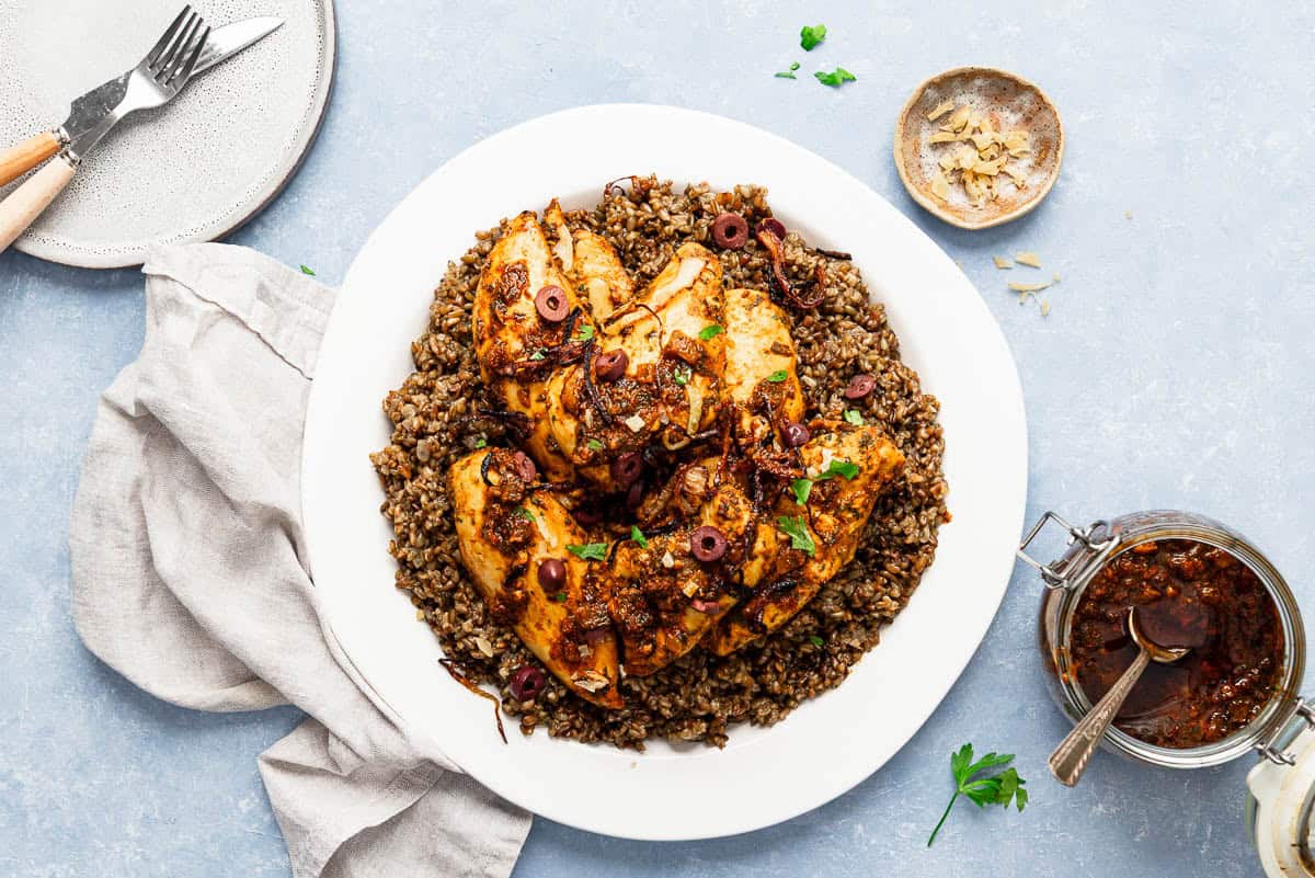 Chicken chermoula on a platter next to a cloth napkin, a plate with a knife and fork, a bowl of the chopped preserved lemon rind, and a jar of red chermoula with a spoon.