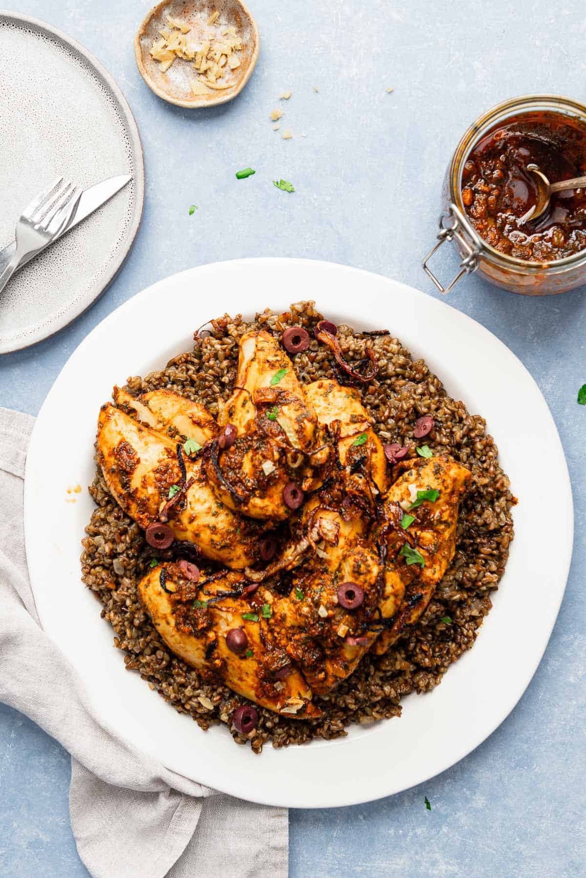 Chicken chermoula on a platter next to a cloth napkin, a plate with a knife and fork, a bowl of the chopped preserved lemon rind, and a jar of red chermoula with a spoon.
