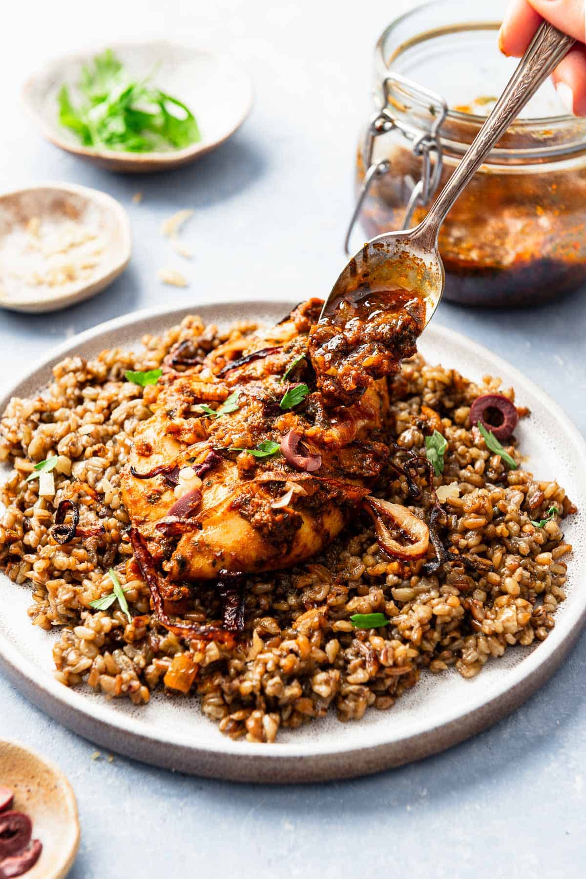 A close up of the red chermoula being spooned onto a serving of the chicken chermoula on a plate. In the background is a jar of the red chermoula, and small bowls of bowls of the chopped preserved lemon rind, sliced kalamata olives and cilantro.